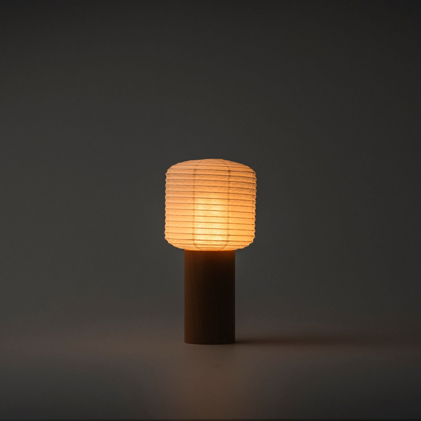 A portable paper table lamp brightly emitting warm light in a dark room, with a wooden handle and paper top