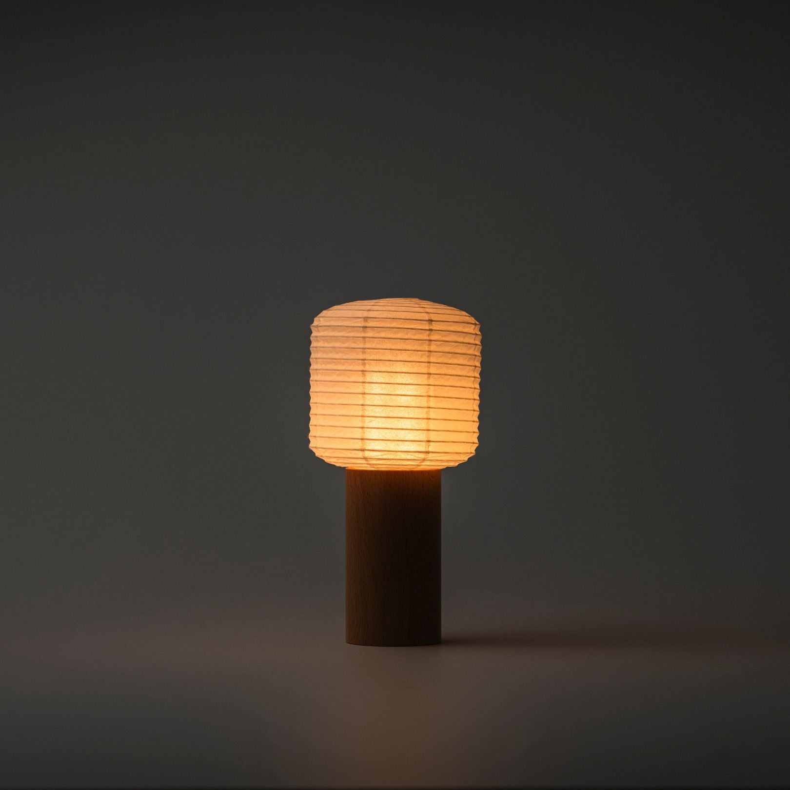 A portable paper table lamp brightly emitting warm light in a dark room, with a wooden handle and paper top