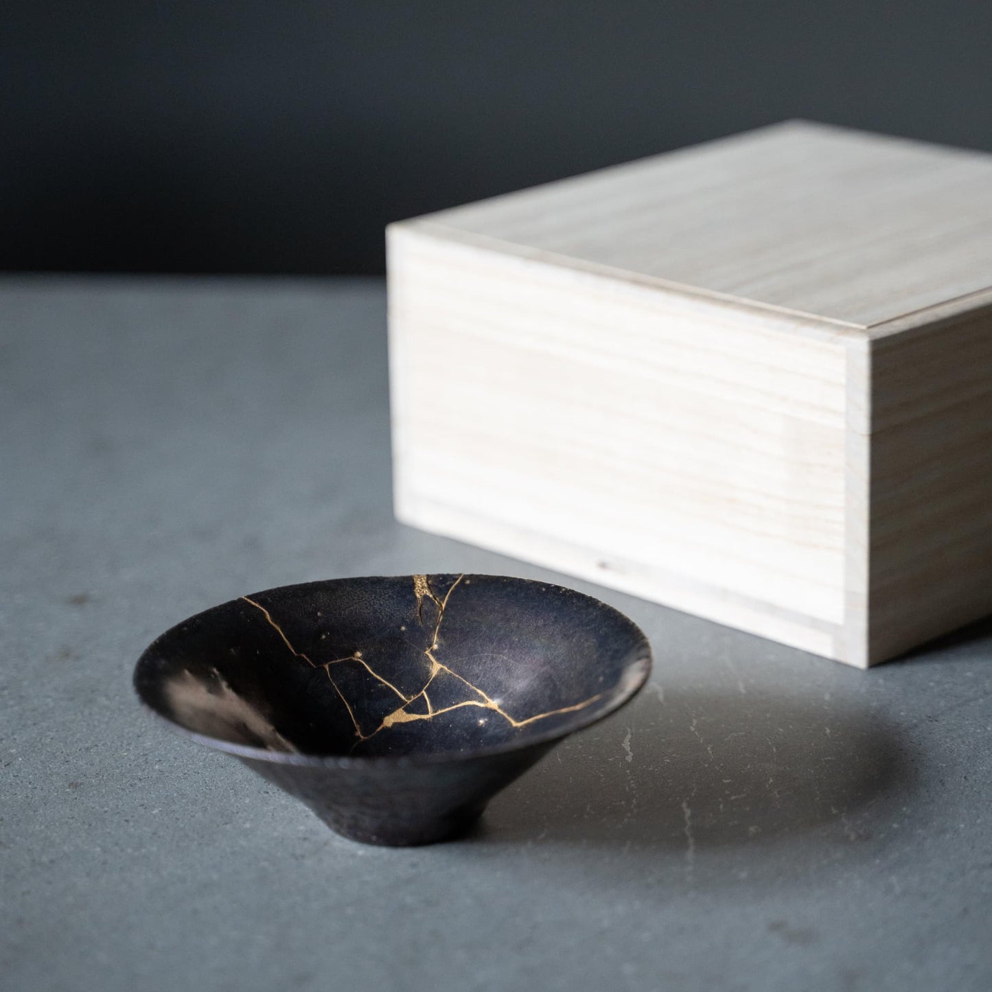 Japandi-style table setting with a grey table featuring a purple bowl repaired with Kintsugi, a traditional Japanese technique, and a paulownia wood box.
