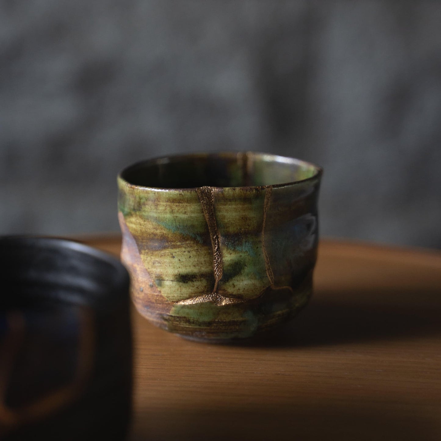 Deep green tea bowl repaired with Kintsugi, a traditional Japanese technique, showcasing the beauty of imperfection, placed on a Japanese-style wooden table.