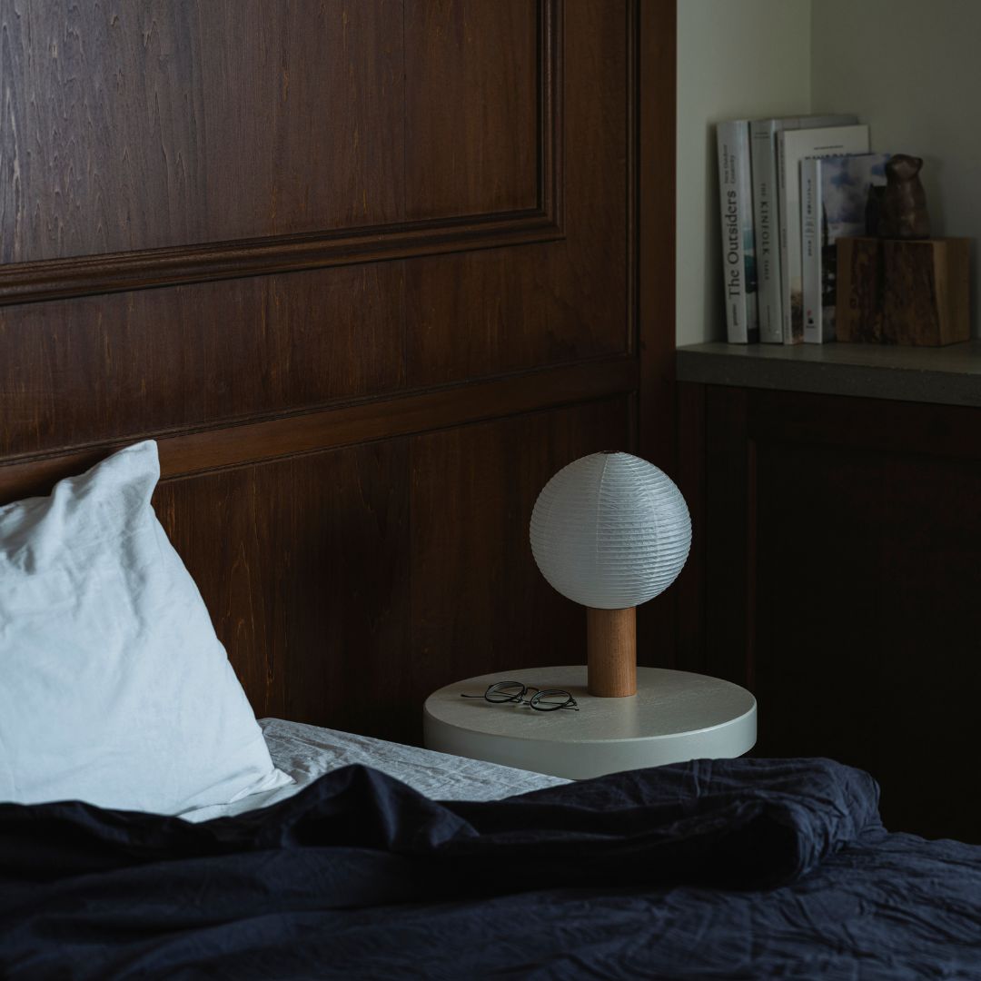 A bedroom with a bed on the left and a bookshelf with books on the right. On the bedside table to the right of the bed is a bedside lamp "TORCHIN" made from Japanese Yame-chochin lanterns.