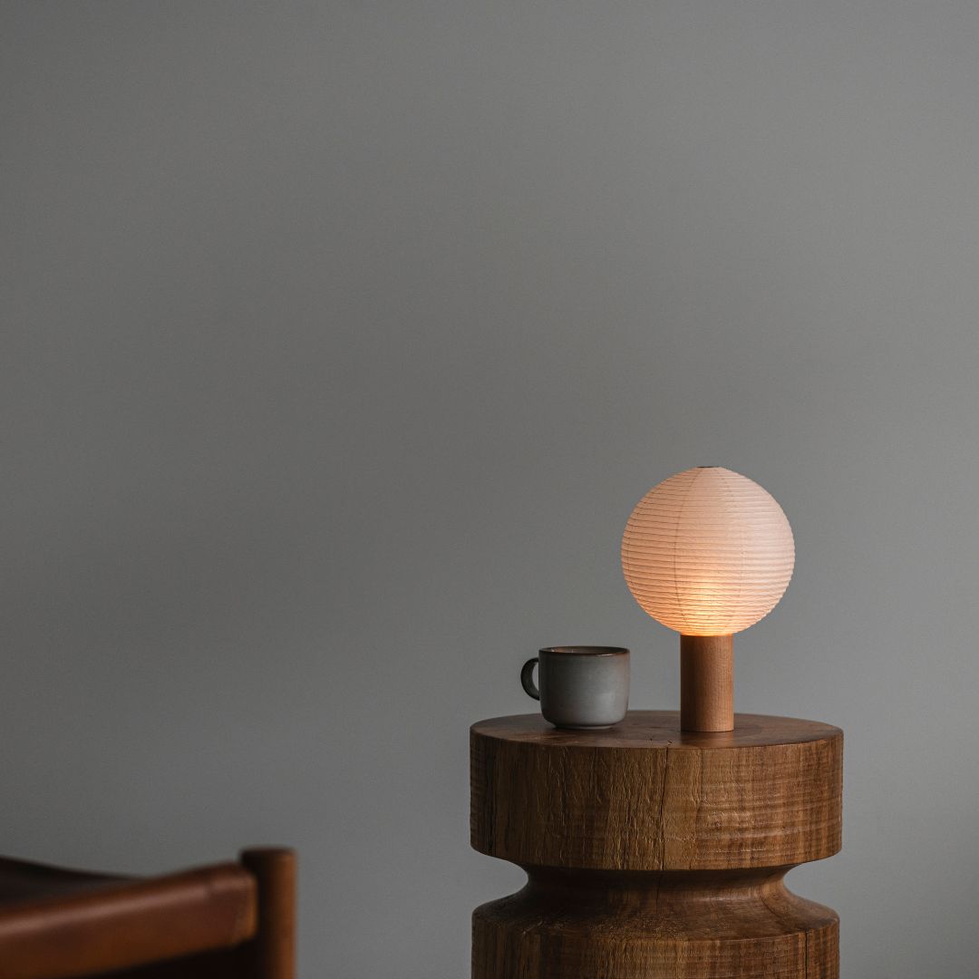 "In een kamer met grijs behang, een houten stoelleuning aan de linkerkant, een ronde houten tafel in het midden-rechts met een washi papieren tafellamp en een mok erop. De draagbare lamp brandt en straalt een warme gloed uit."