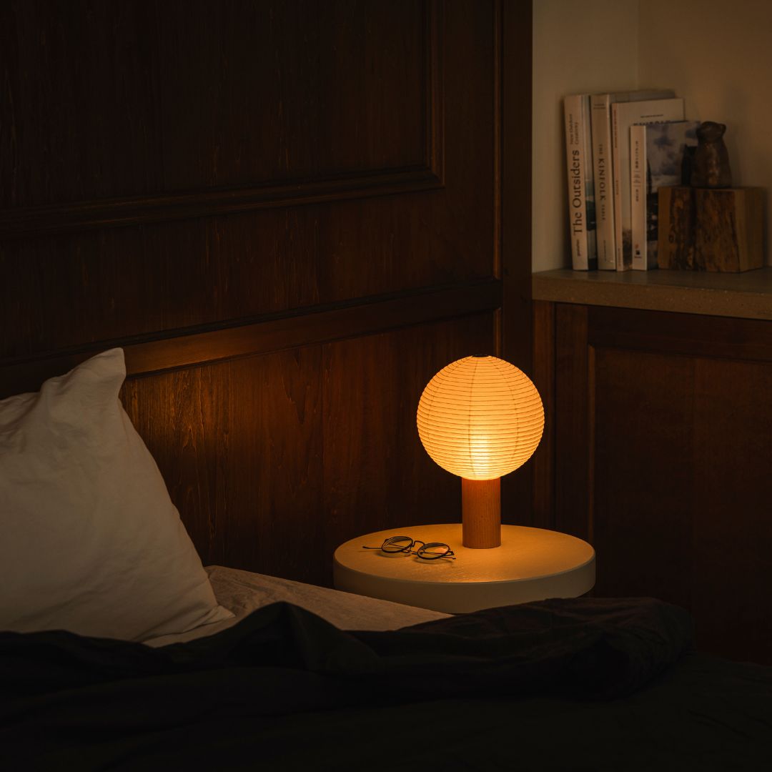 "Een schemerige slaapkamer met links een bed en rechts een boekenplank met boeken. Op het nachtkastje rechts van het bed staat een nachtlampje \"TORCHIN\" gemaakt van Japanse Yame-chochin lantaarns, dat een warme gloed uitstraalt vanuit de sferische kap."