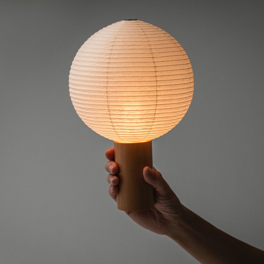 Against a gray wall, a portable lamp "TORCHIN" made using the techniques of Yame-chochin lanterns from Yame, Fukuoka, Japan, is lit in the center, emitting a warm lamp. The lampshade is a round lantern, and it has a wooden cylindrical handle being held by a hand.