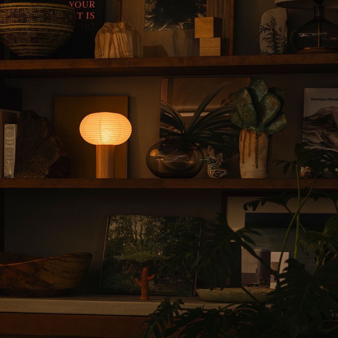 A dimly lit room with a three-tier wooden shelf against the back wall, decorated with various items. On the left side of the second shelf is a portable lamp "TORCHIN" made from Japanese Yame-chochin lanterns, emitting a warm lamp from its oval shade.