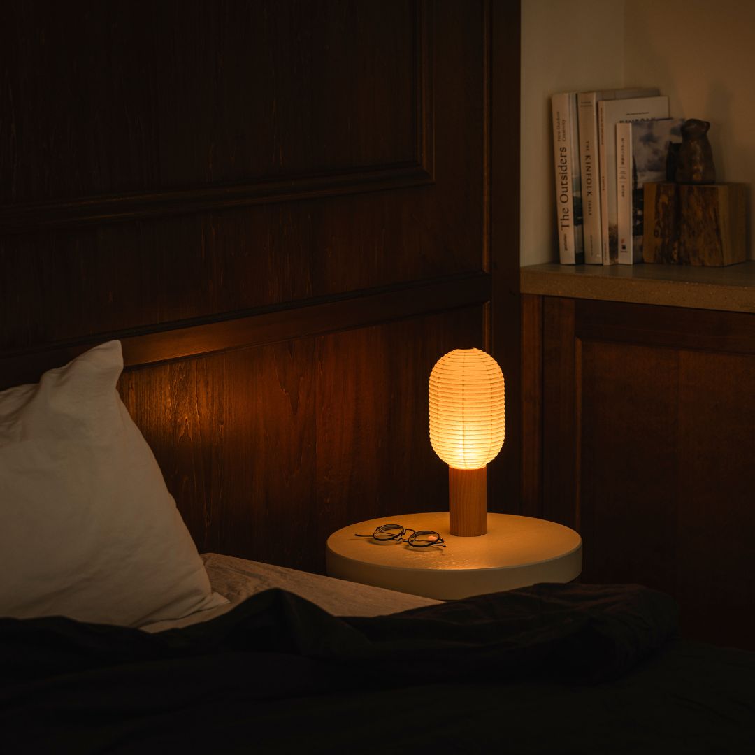 A bedroom with a bed on the left and a bookshelf with books on the right. On the bedside table to the right of the bed is a bedside paper lamp "TORCHIN" made from Japanese lanterns, emitting a warm lamp from its cylindrical shade.