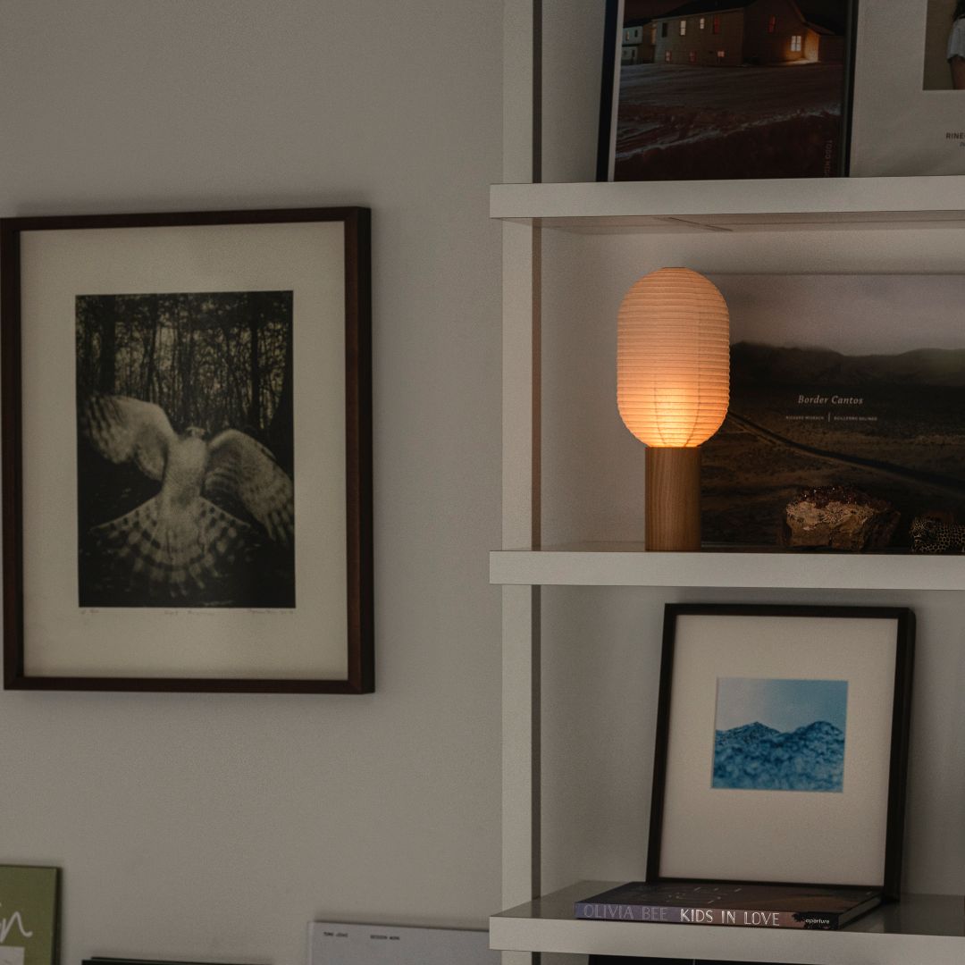A bright white room with a wall art on the left and a white three-tier bookshelf on the right. Two portable lamps "TORCHIN" are displayed on the second shelf, emitting a warm lamp from their spherical shades.