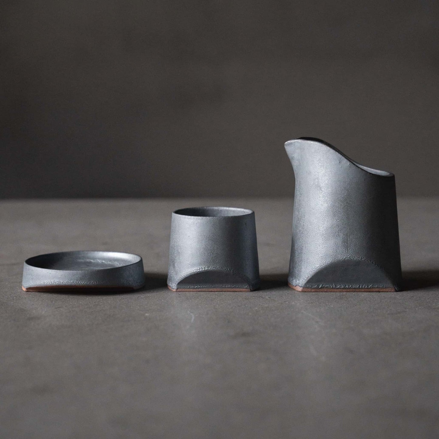Iron-colored sake pitcher, cups, and plate, crafted using traditional Japanese roof tile techniques, arranged on a grey table.