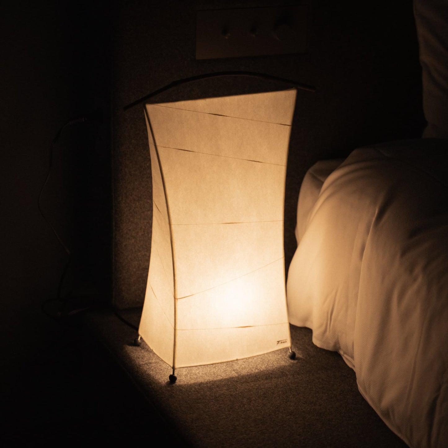 A Japanese paper lamp casts a warm glow beside a bed in a dimly lit bedroom