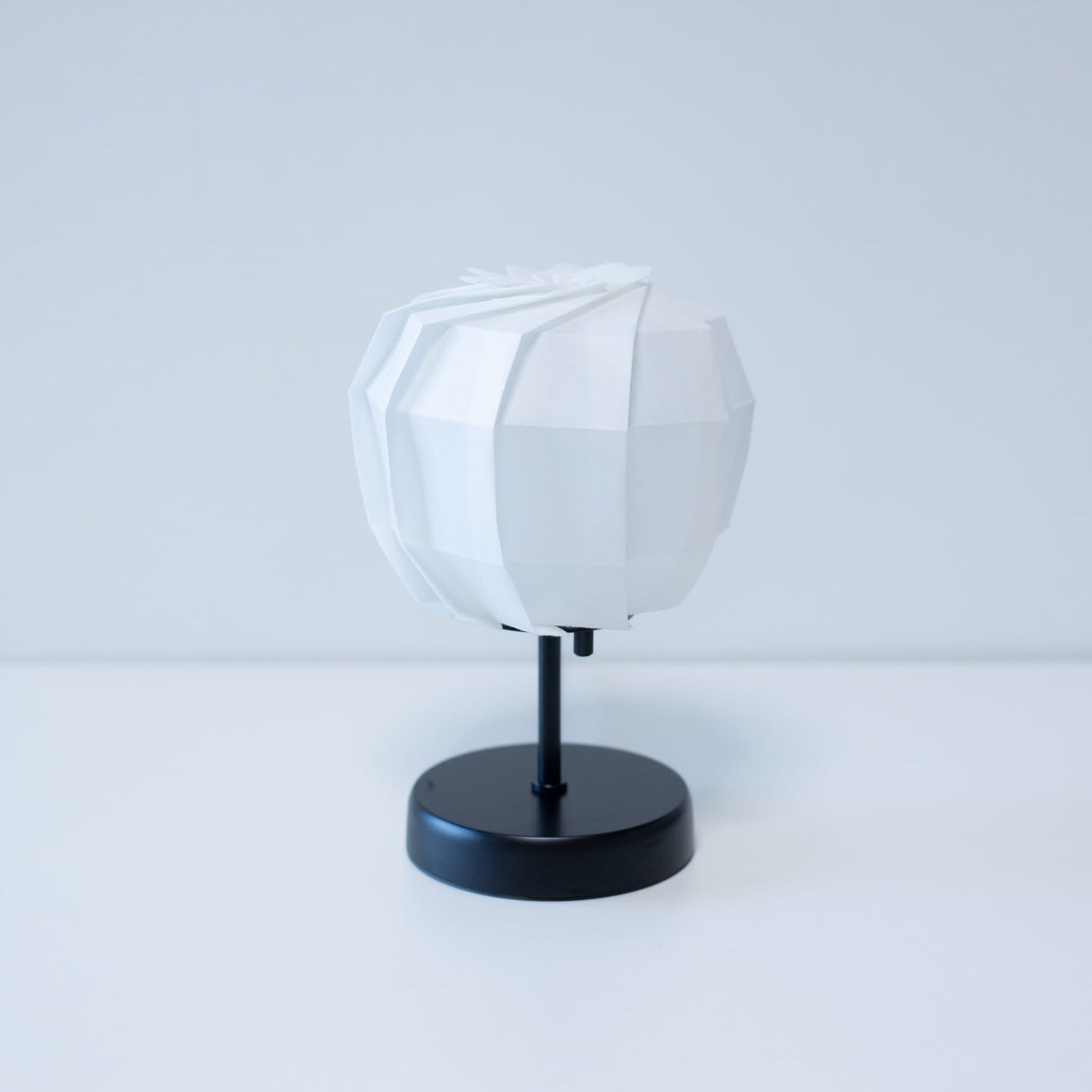 Stylish Origami table lamp featuring a black stand and a spherical white lampshade crafted from intricately folded washi paper.
