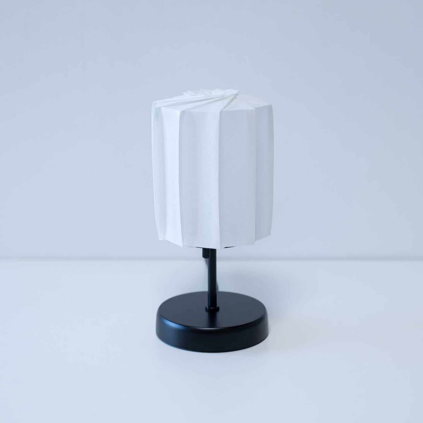 Origami table lamp "tutu" with a black stand and a white cylindrical lampshade made of pleated washi paper, adding an elegant touch to any room.