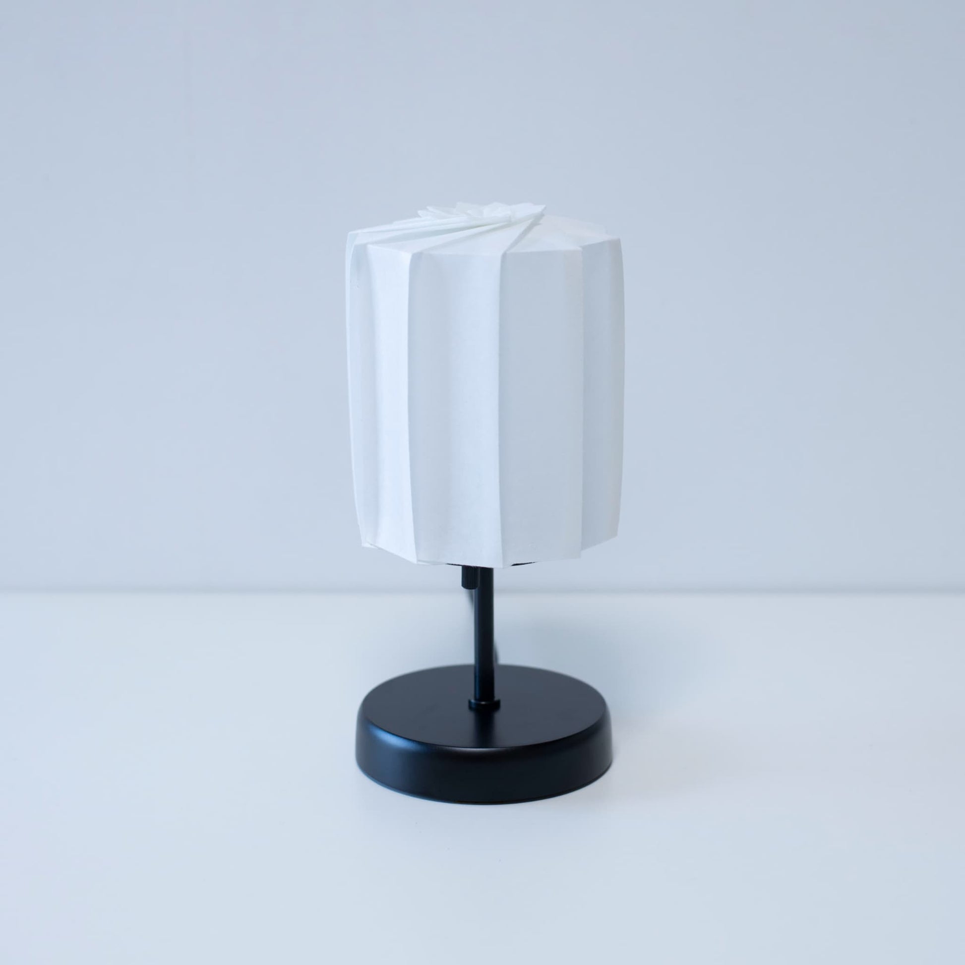 Origami table lamp "tutu" with a black stand and a white cylindrical lampshade made of pleated washi paper, adding an elegant touch to any room.