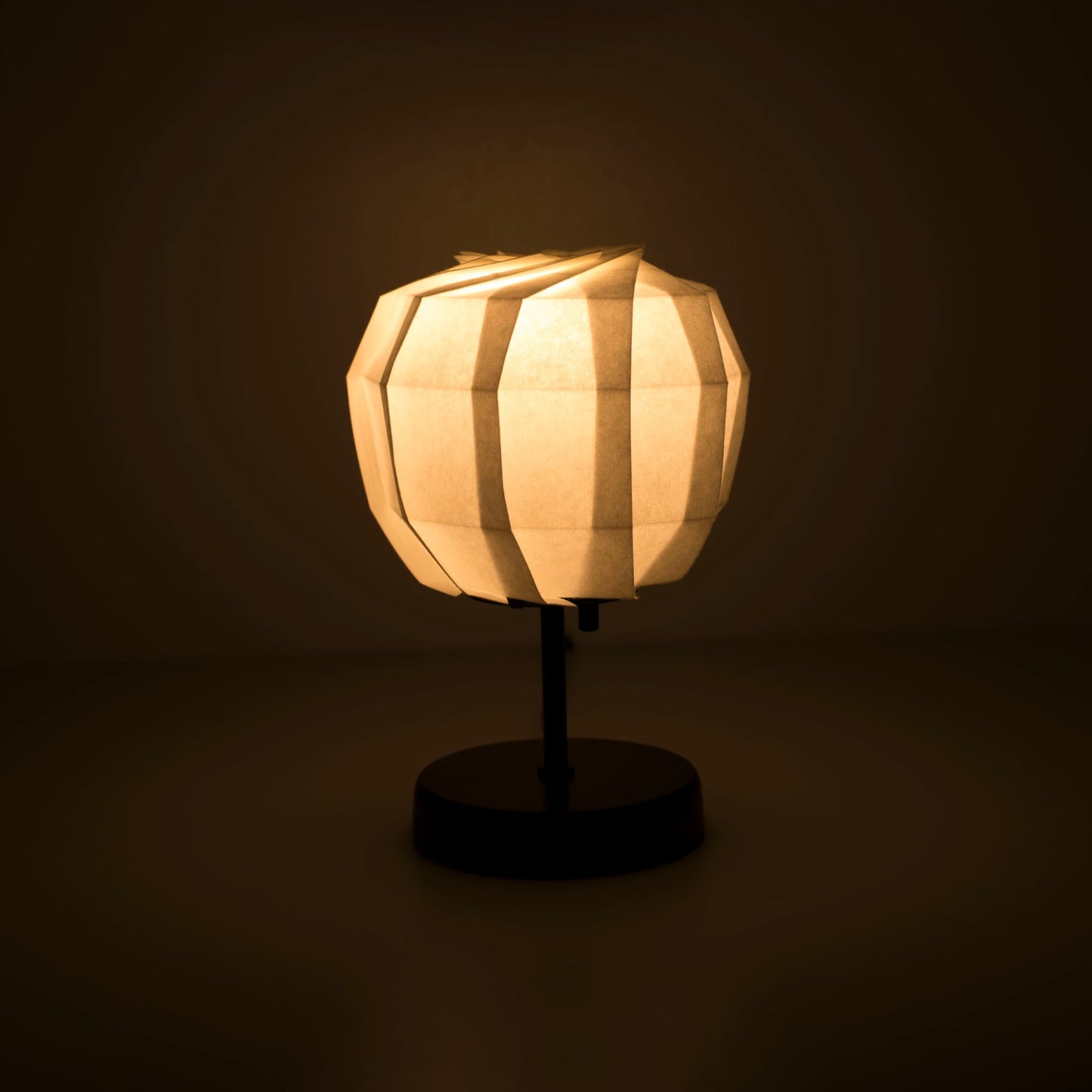Origami paper table lamp "maru" with a black stand and a spherical white lampshade crafted from intricately folded washi paper, emitting a warm and inviting light.