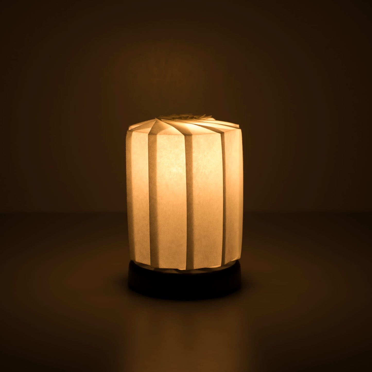 Origami Table lamp "tutu M" with a black base and a cylindrical white lampshade made of pleated washi paper, emitting a warm and inviting light.