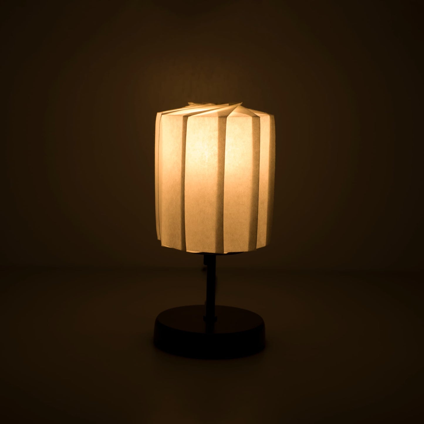 Origami paper table lamp "tutu" with a black stand and a cylindrical white lampshade made of pleated washi paper, casting a soft and warm light.