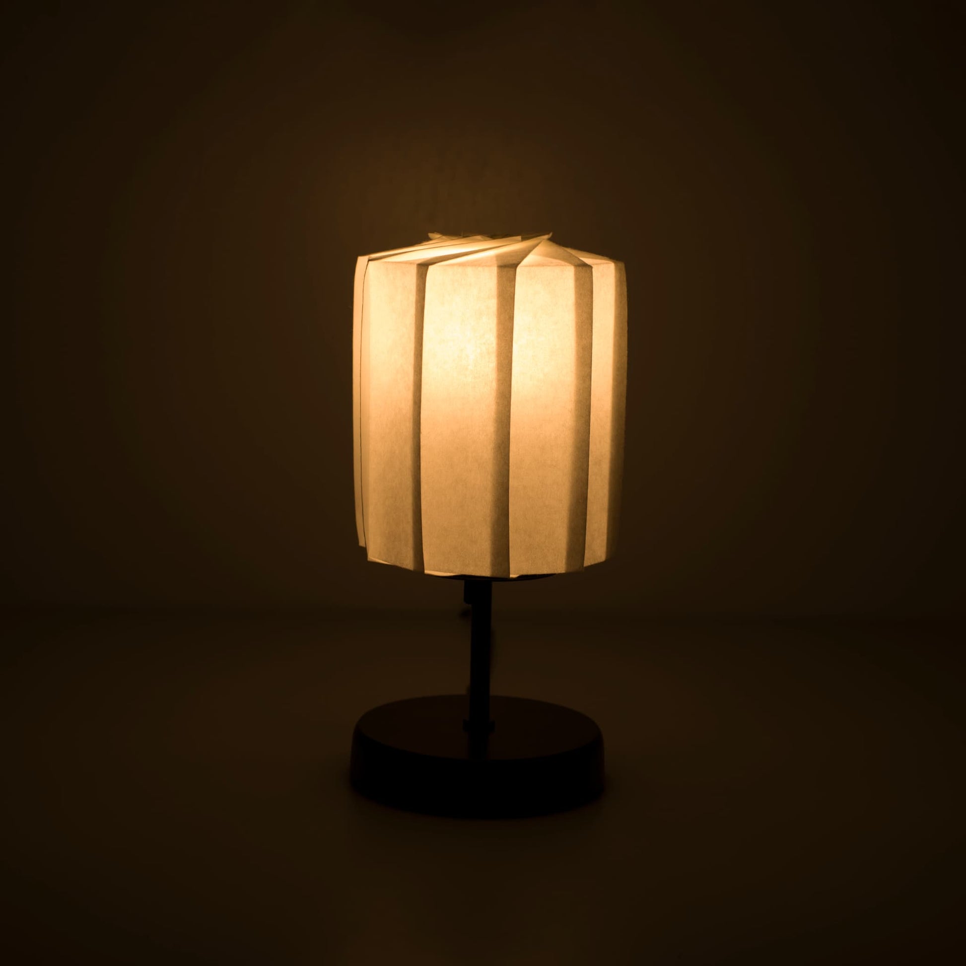 Origami paper table lamp "tutu" with a black stand and a cylindrical white lampshade made of pleated washi paper, casting a soft and warm light.