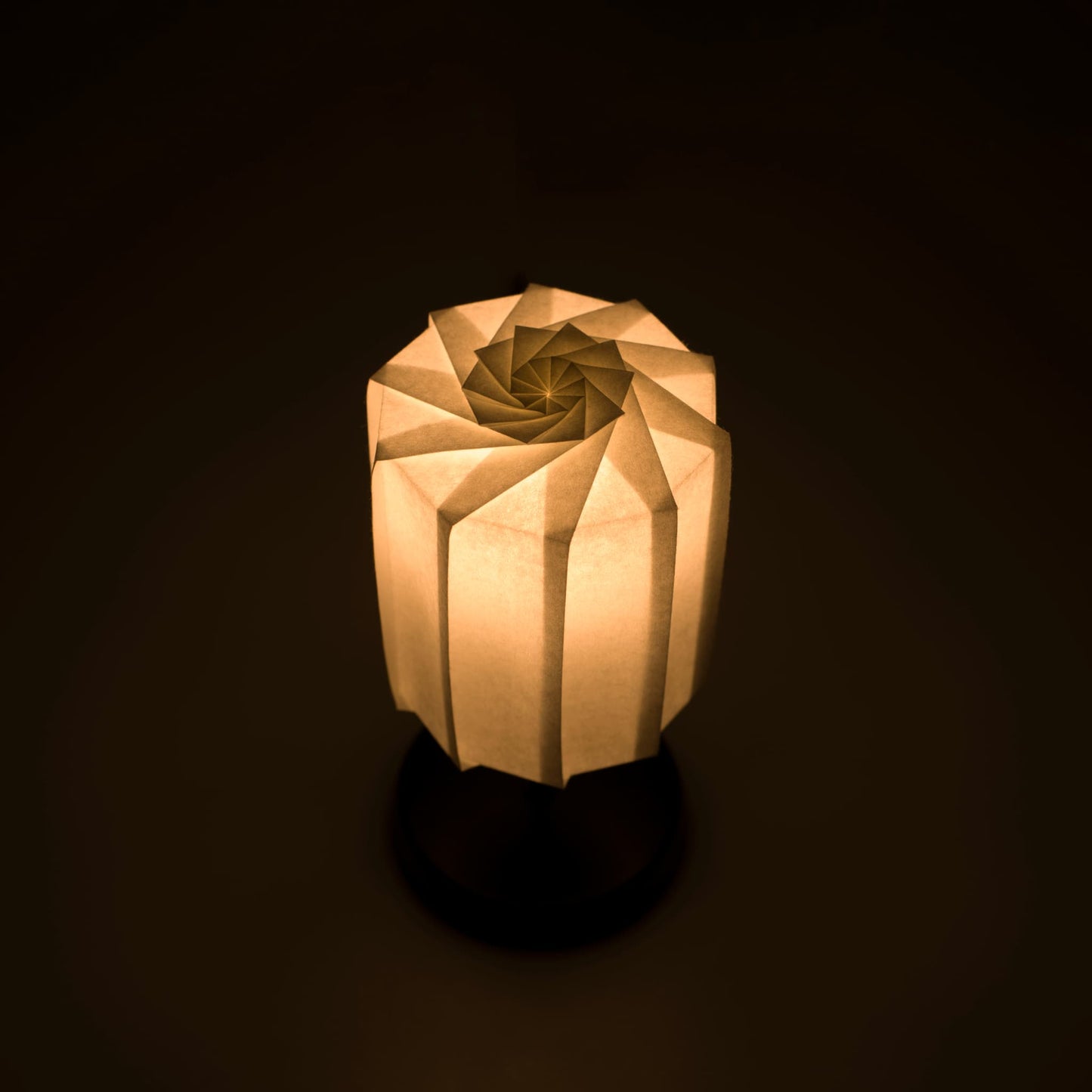 Origami paper table "tutu" lamp with a black stand and a cylindrical white lampshade made of pleated washi paper, emitting a warm and inviting light.