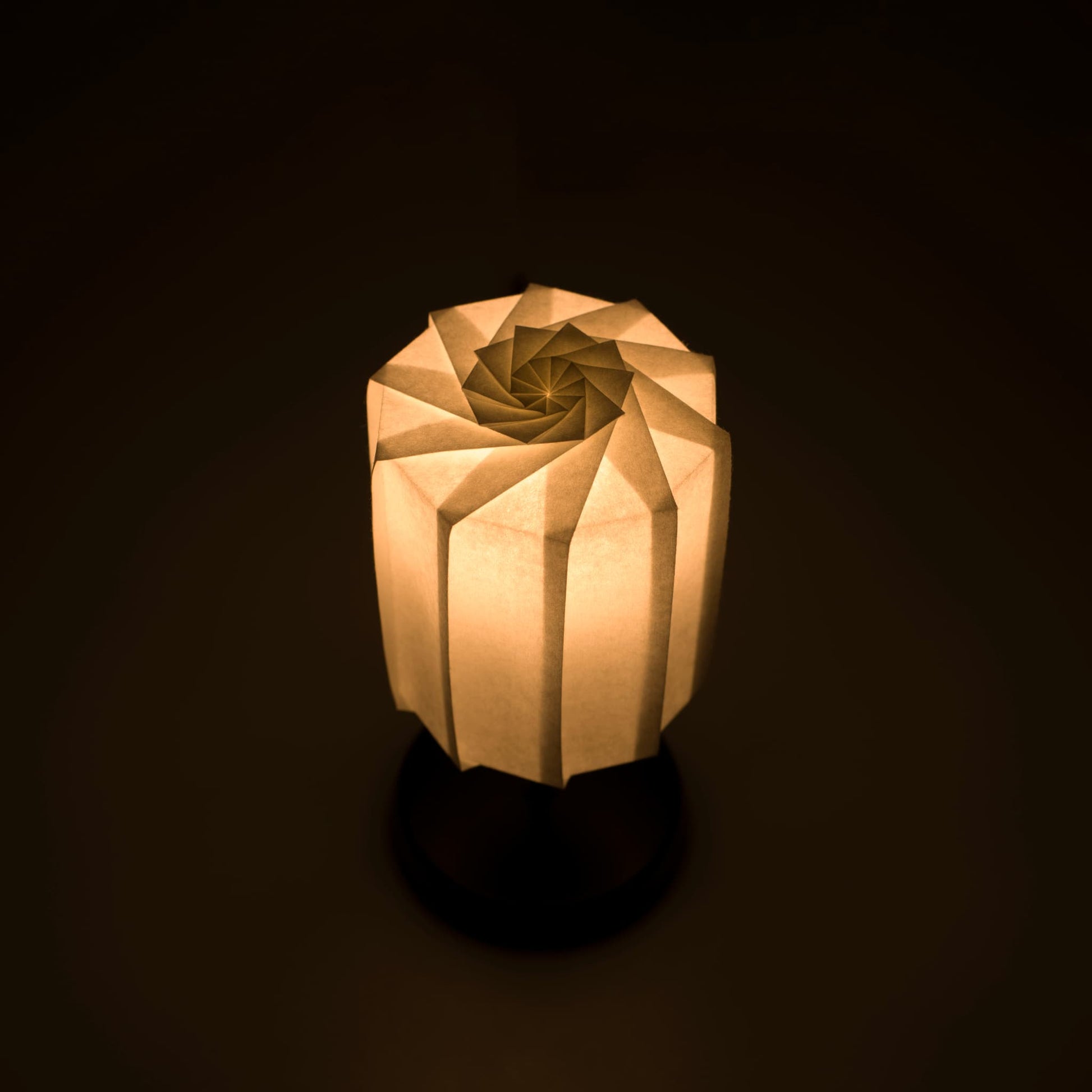 Origami paper table "tutu" lamp with a black stand and a cylindrical white lampshade made of pleated washi paper, emitting a warm and inviting light.