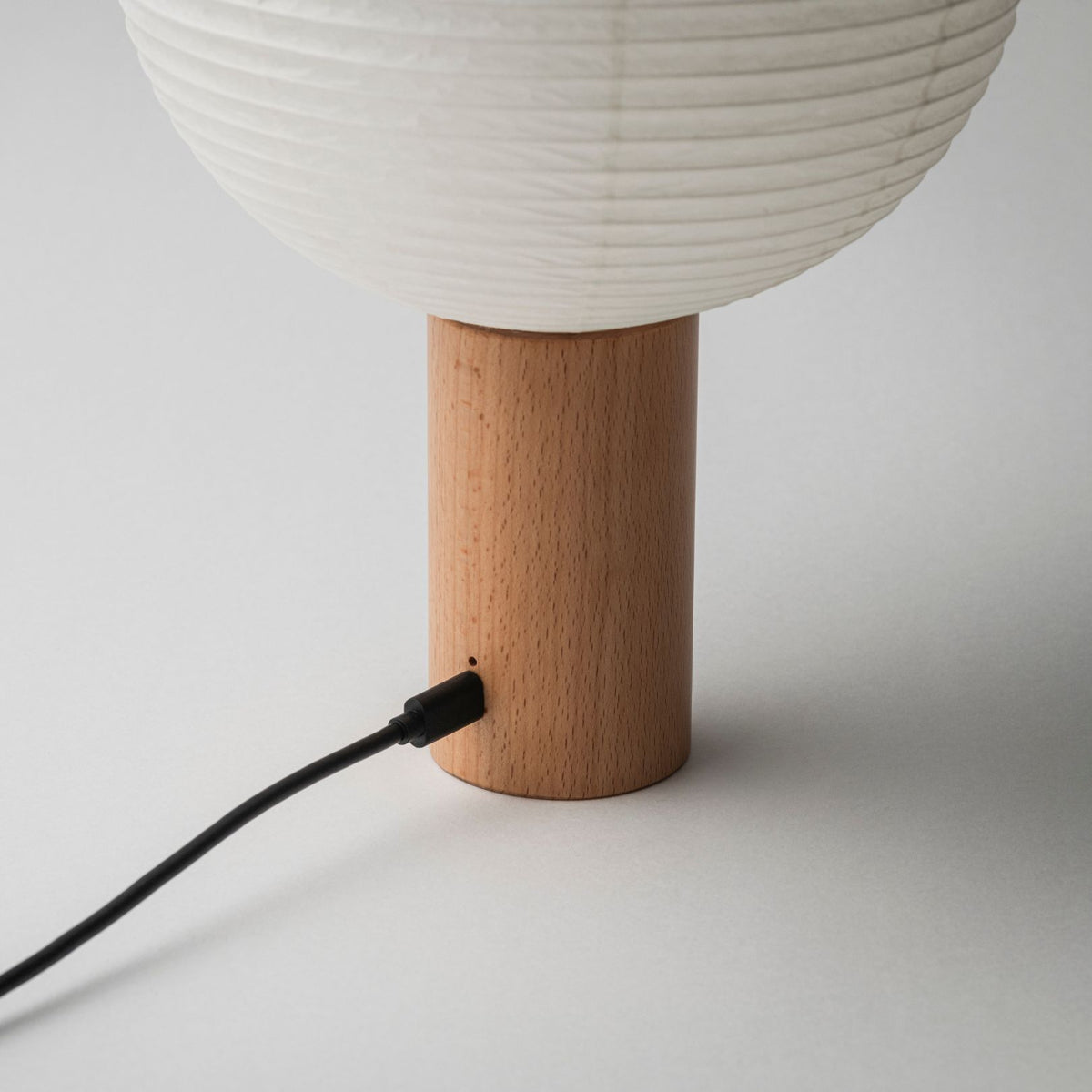 "Een draagbare papieren lamp met een houten voet, voorzien van een oplaadpoort, aangesloten op een oplaadkabel"