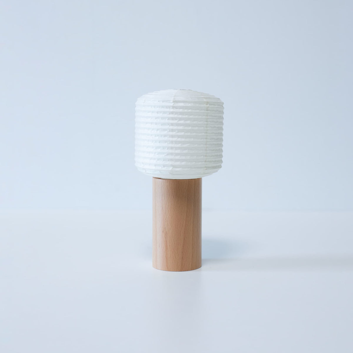 Een draagbare papieren lamp met een houten voet en een cilindrische papieren kap tegen een witte achtergrond