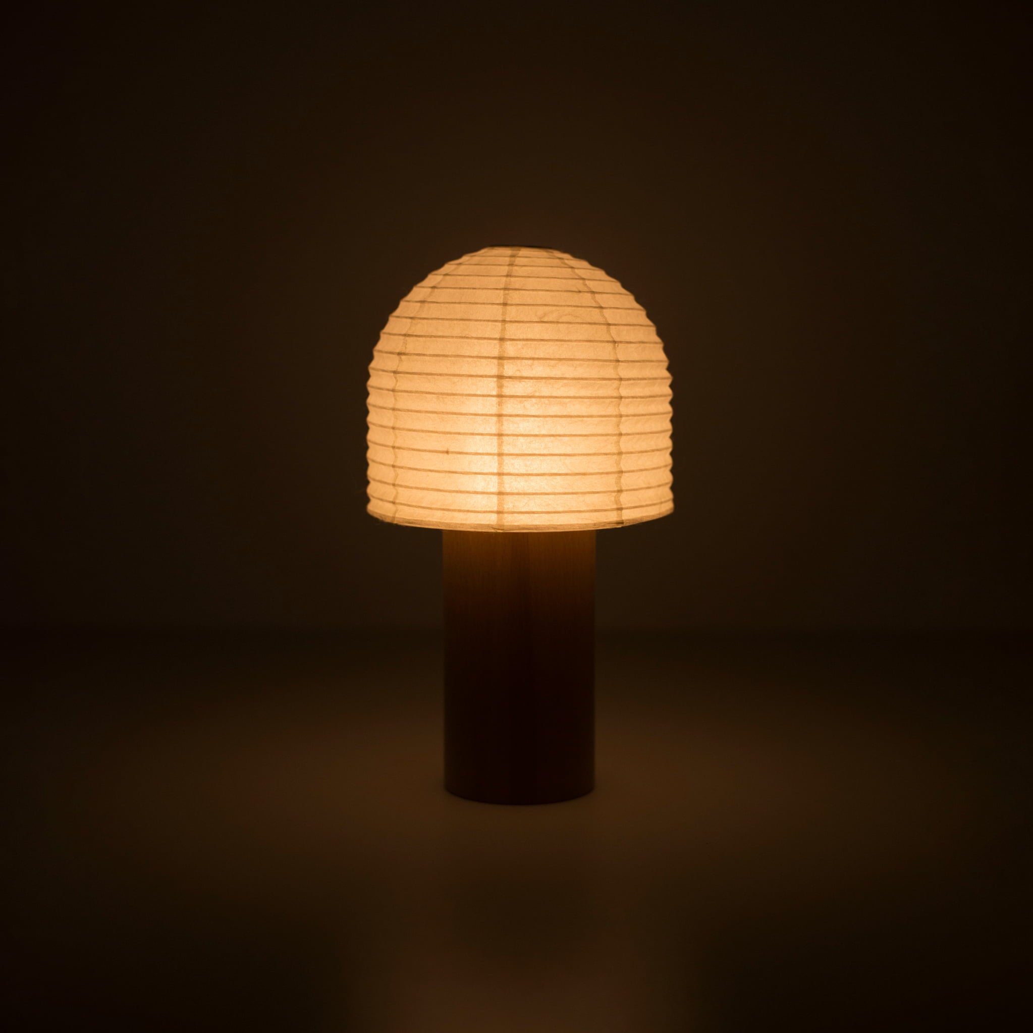 A portable paper lamp with a wooden handle and bell-shaped cylindrical paper top emitting warm light in a dark room