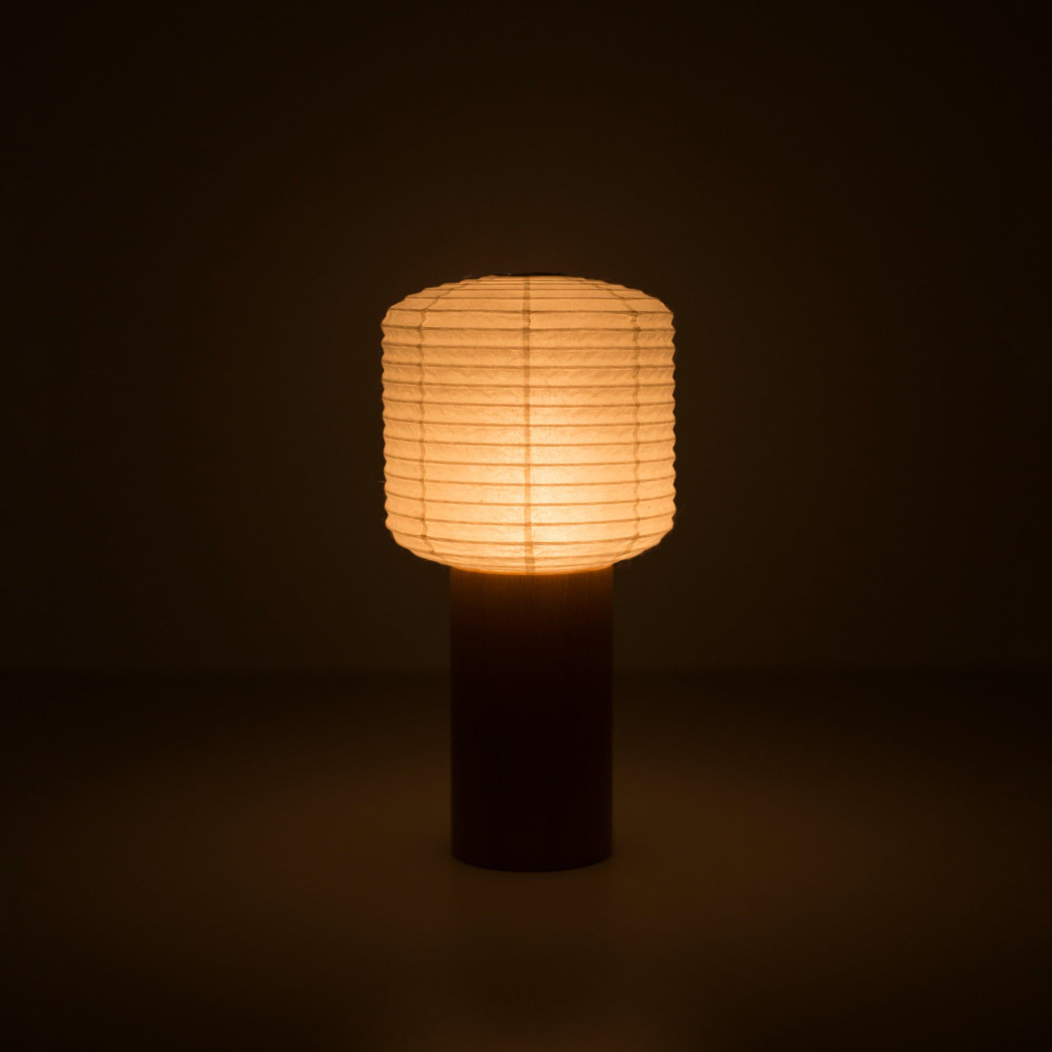 A portable Japanese paper lamp with a wooden handle and square-shaped cylindrical paper top emitting warm light in a dark room