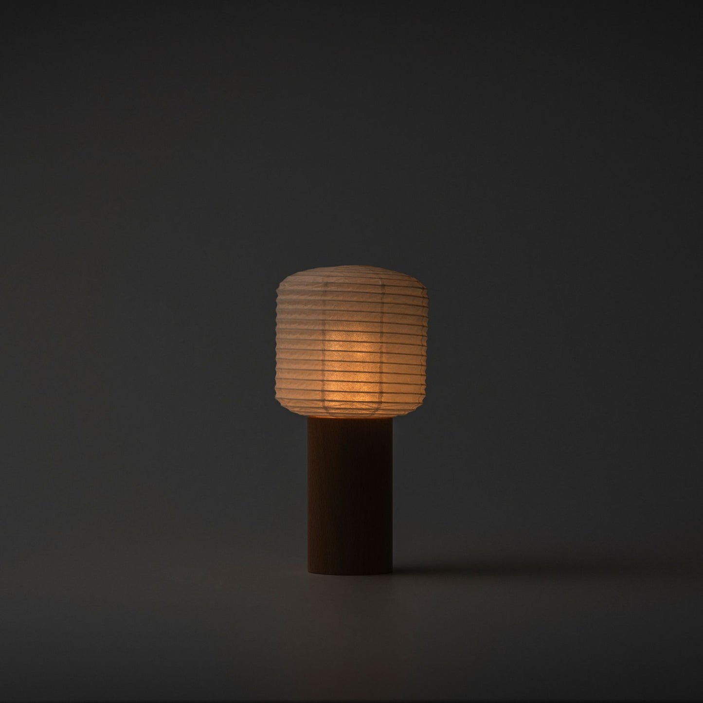 A portable paper lamp softly glowing with a warm light in a dark room, featuring a wooden handle and paper top
