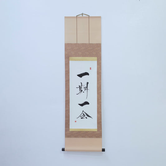 Brown hanging scroll with Japanese calligraphy of '一期一会' (one-time, one meeting) on white rice paper, mounted on a white wall