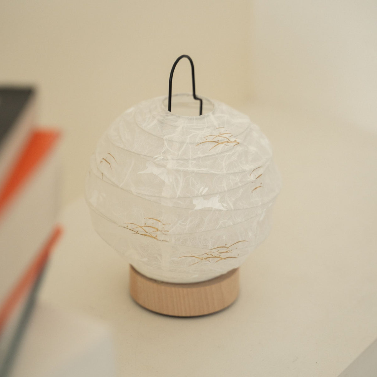 Close-up van een onverlichte, ronde Japanse papieren tafellamp "Yuhi" met een konijn, die het getextureerde papieren oppervlak toont voor wabi-sabi decoratie op een Japandi-woonkamerconsole.