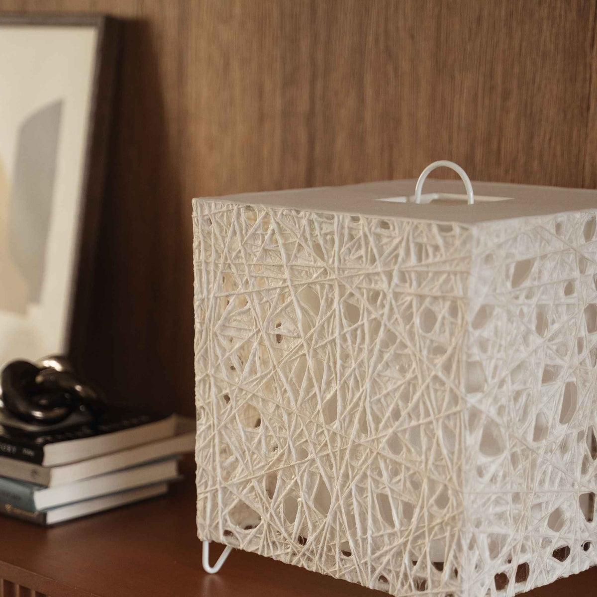 White cube shaped Japanese paper lamp Me Look 03 sitting unlit on a wooden console table near a bed background.