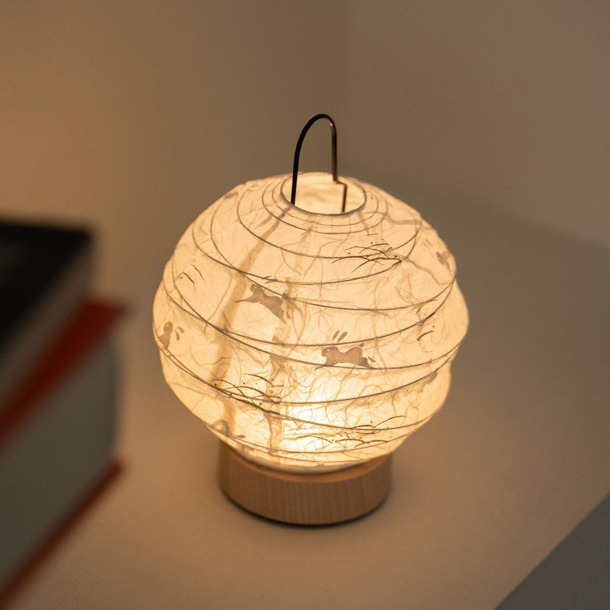 Glowende ronde Japanse papieren tafellamp "Yuhi" Konijn dient als een gezellige accentlamp naast boeken op een witte console tafel.