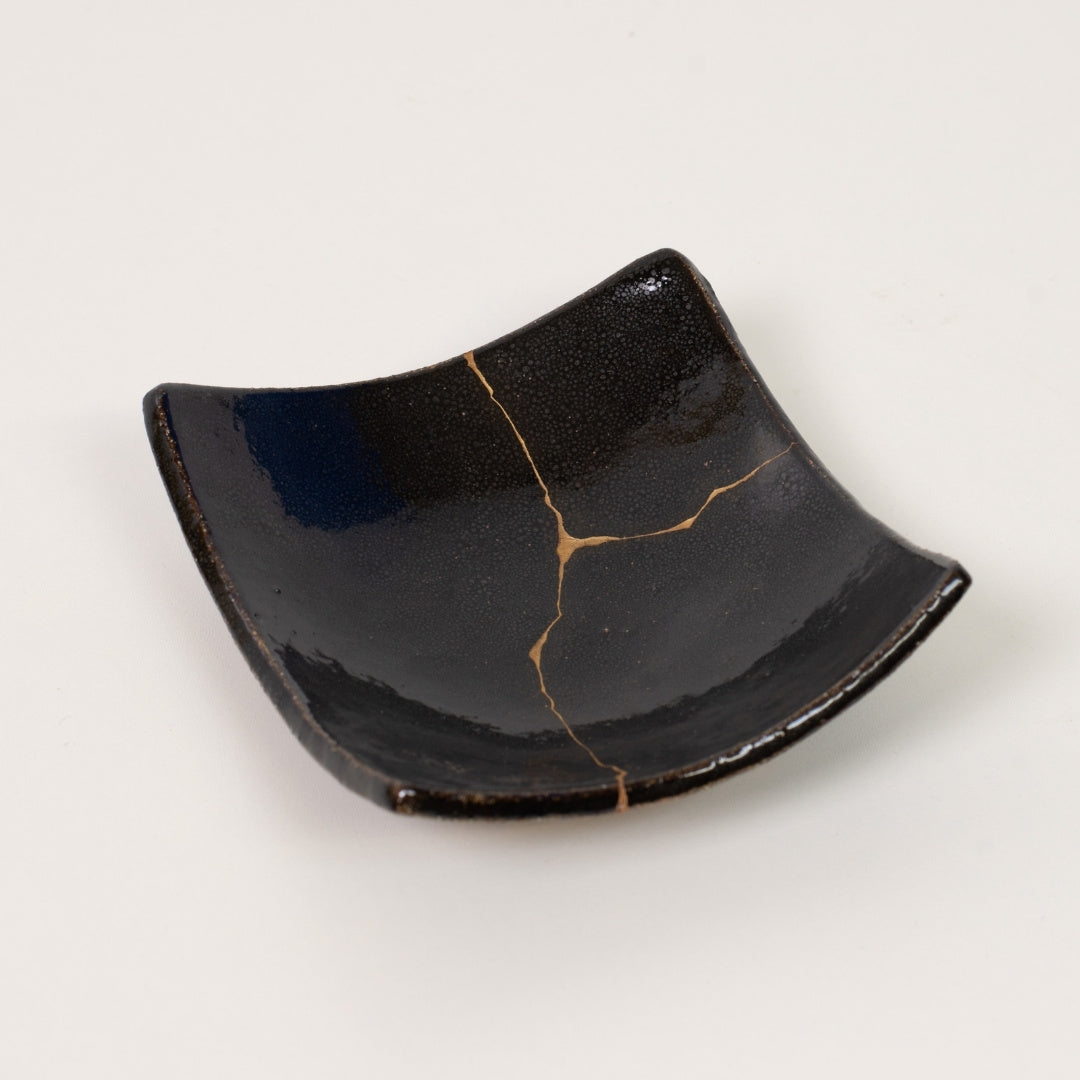 A sophisticated black square kintsugi plate rests directly on a white surface highlighting the authentic gold repair
