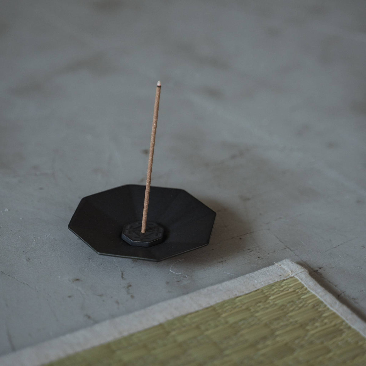 "Assiette à encens japonaise noire octogonale en porcelaine d'Arita avec un bâtonnet allumé près d'un tapis tatami."
