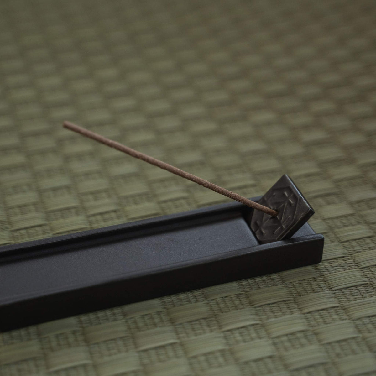 Support à encens japonais rectangulaire sombre sur un tapis tatami avec un bâtonnet allumé créant une atmosphère calme.