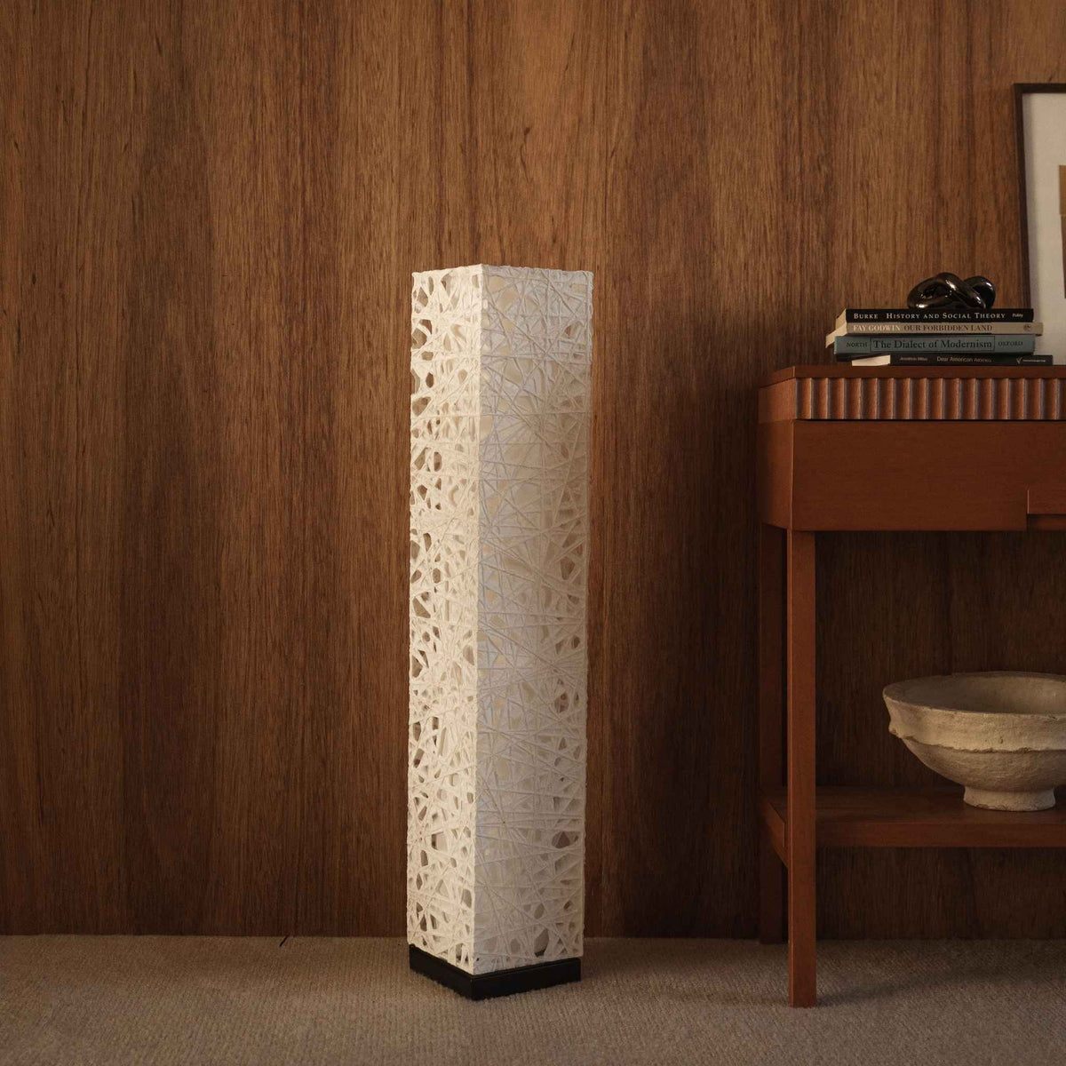 Tall white Me Look 01 Japanese paper lamp standing unlit next to a wooden console table in a living room.