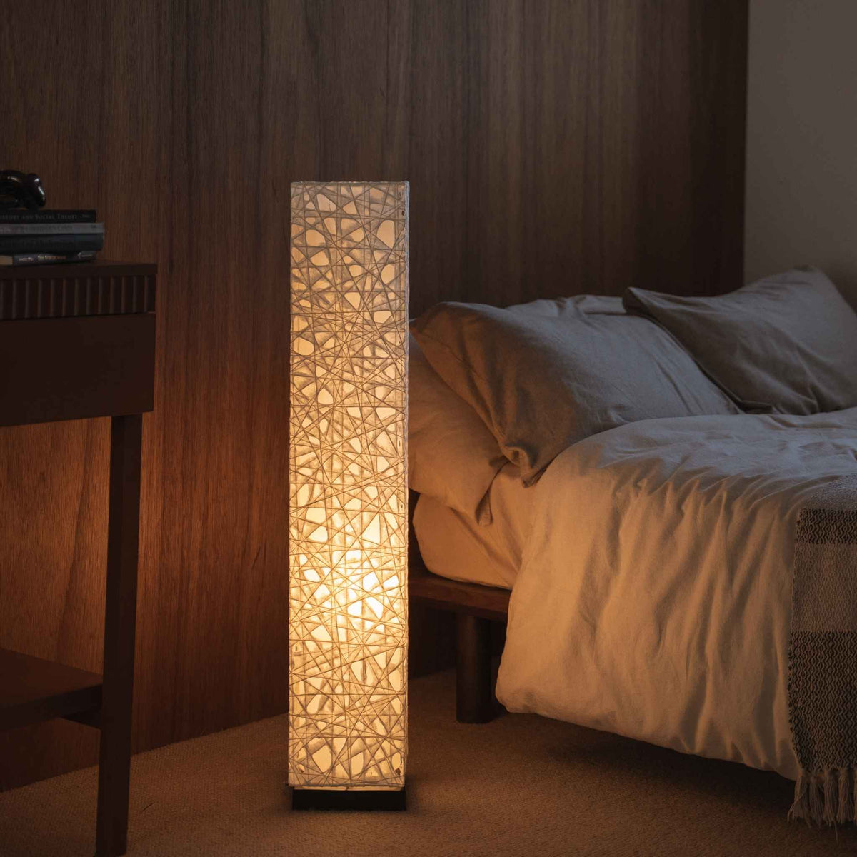 Glowing white Japanese paper floor lamp Me Look 01 providing warm light next to a bed in a japanese bedroom.