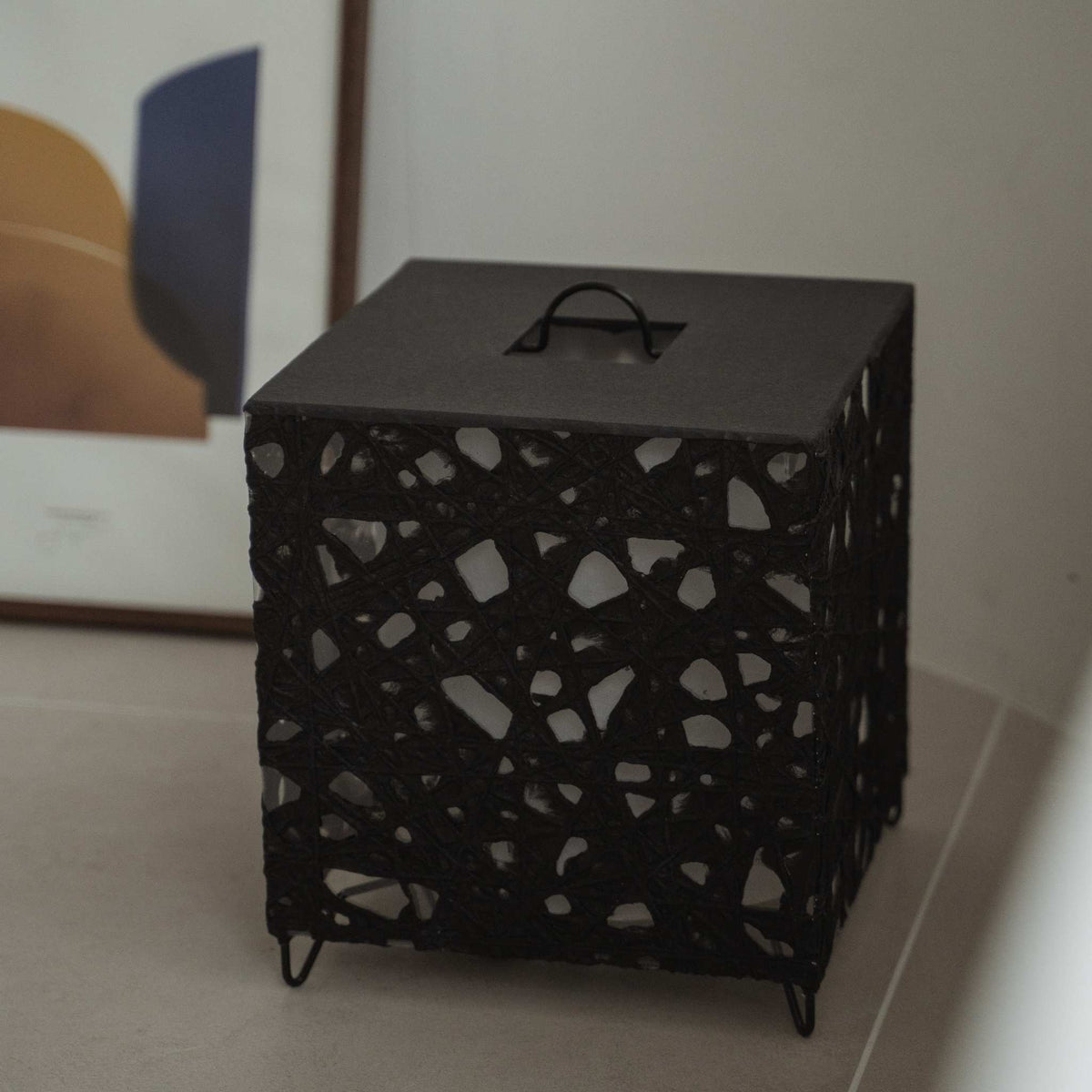Unlit black cube Japanese floor lamp Me Look 03 sitting on a beige tiled floor next to a framed art print