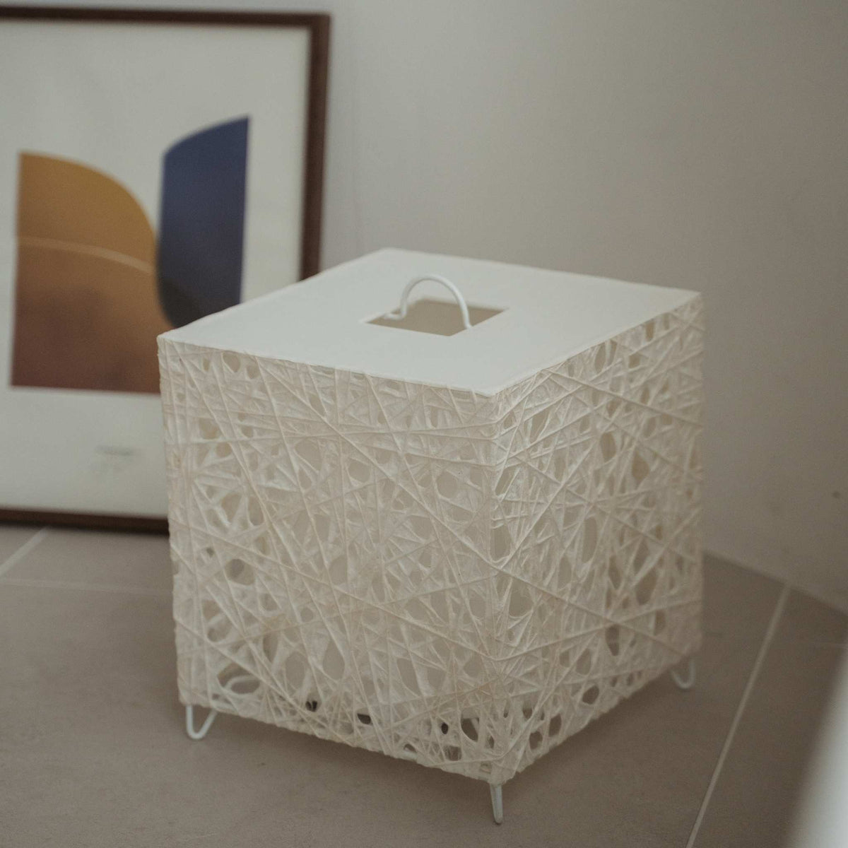 Unlit white cube Japanese floor lamp Me Look 03 sitting on a beige tiled floor next to a framed art print