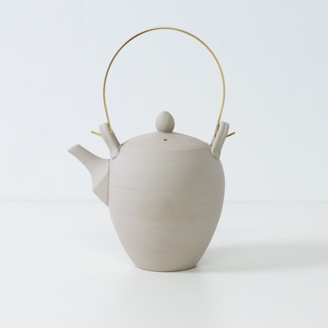 Side view of a donguri white japanese teapot with a brass handle showing the matte unglazed banko yaki stoneware texture