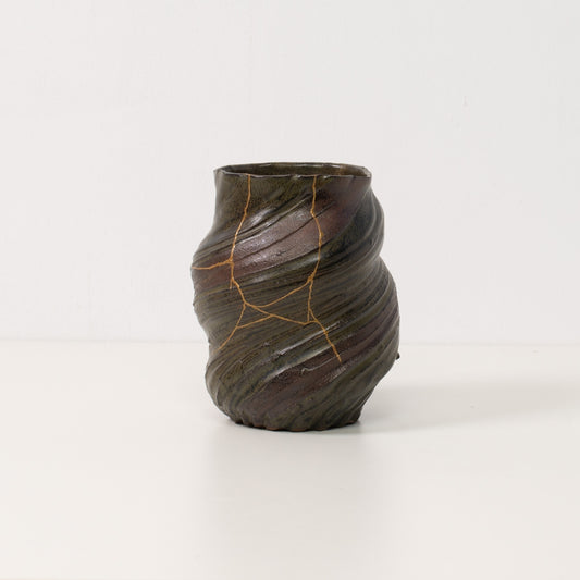 A twisted dark kintsugi vase stands alone on a white table showcasing golden lines of authentic kintsugi pottery art
