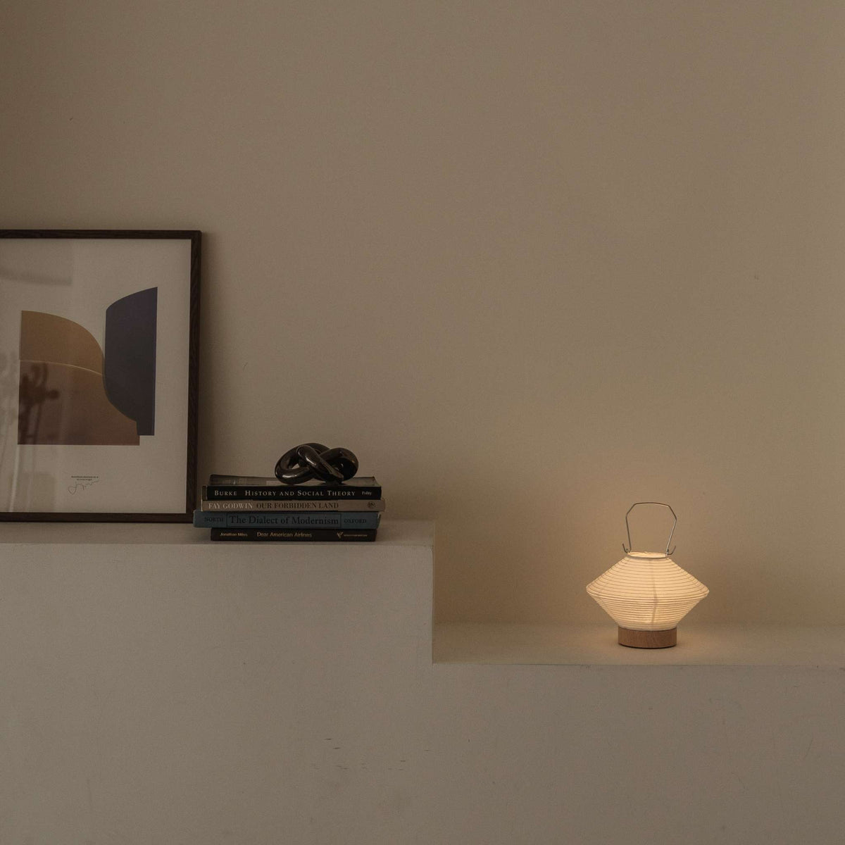 Lysende japansk papir bordlampe RIN 05 på en hylde ved siden af bøger, der giver blødt omgivende lys, velegnet til et Japandi-stil rum