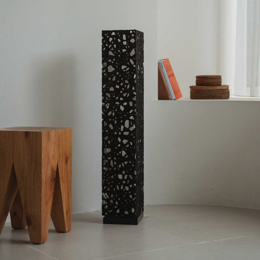 Tall black Japanese paper floor lamp Me Look 01 unlit in a white room next to a wooden stool and books.