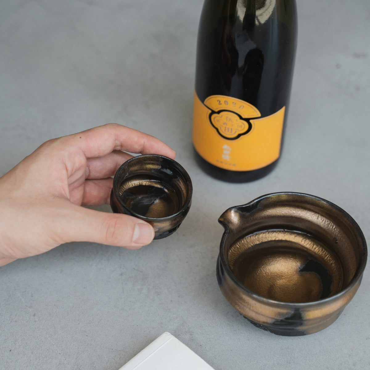 A hand carefully lifting a handcrafted sake cup (ochoko) from a concrete surface to sip, with the sake carafe and bottle nearby.