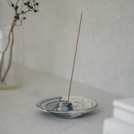 White and grey Japanese incense holder with a lit stick on a light tabletop, creating serenity for a morning meditation.