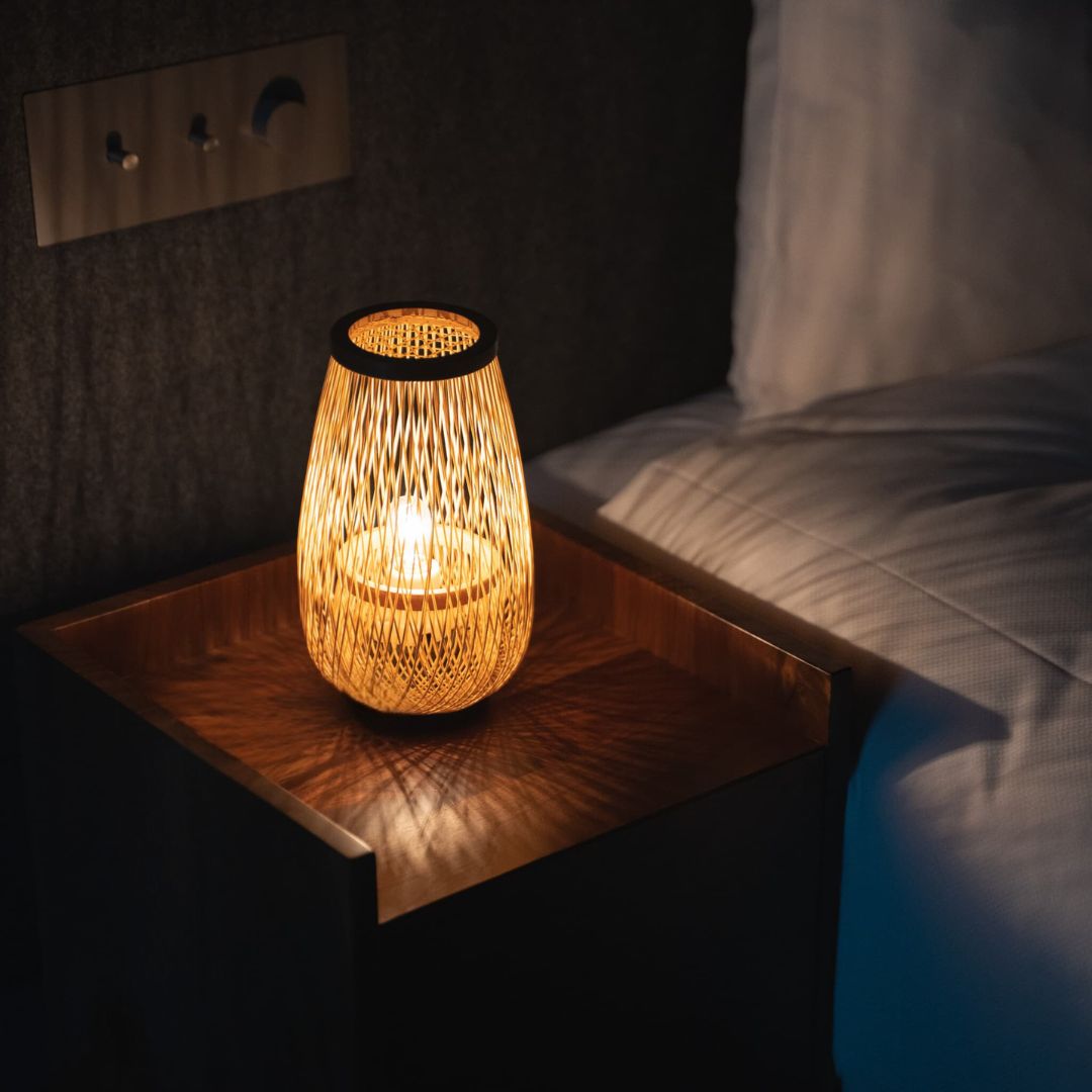 A dimly lit bedroom with a bedside table in the center. A cylindrical Japanese Bamboo Table lamp emits a soft light on the table.