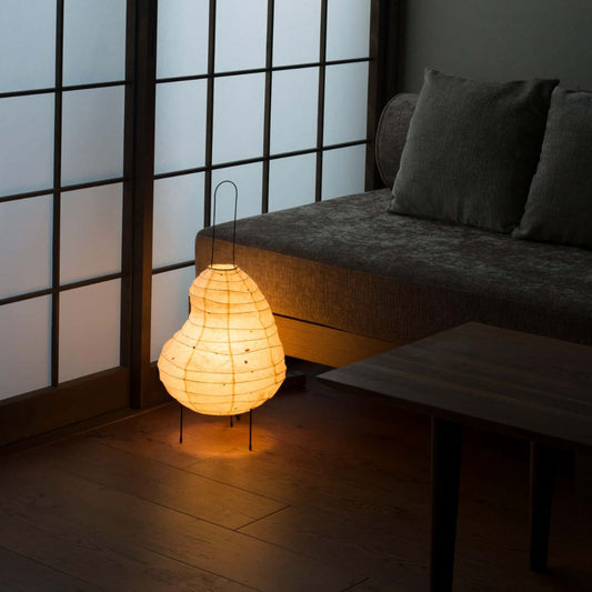 A dimly lit Japanese-style room with shoji screens on the left wall and a gray sofa on the right. A bean-shaped Japanese paper floor lamp, "Beans light," emits a warm light in the center of the photo.