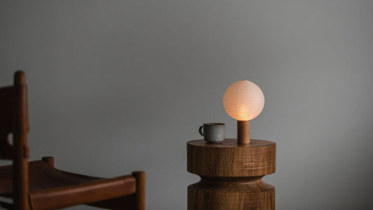 In a room with gray wallpaper, a wooden chair armrest on the left, a round wooden table in the center-right with a washi paper table lamp and a mug on it. The portable lamp is lit, emitting a warm lamp.