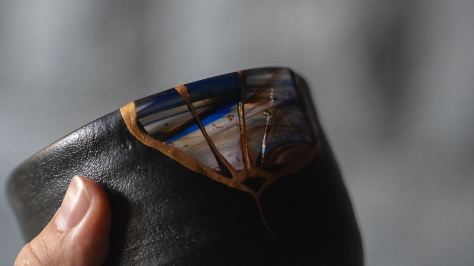A hand holds a jet black Kintsugi tea bowl Orion showcasing gold lines and blue glass in a wabi-sabi pottery style.