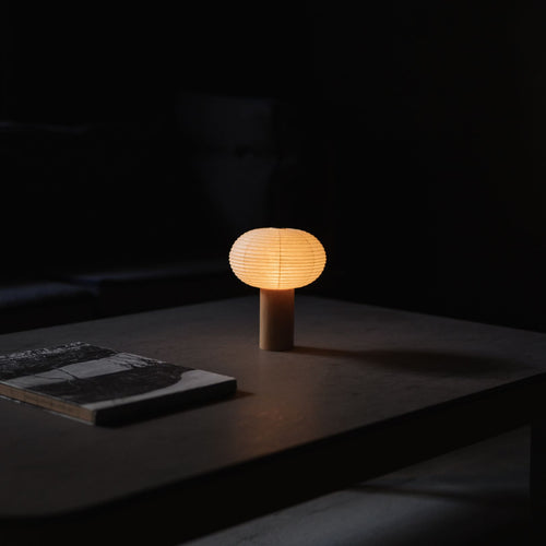 Japanese Paper Table Lamp "TORCHIN OVAL" emitting warm light on a gray table in a dimly lit room.