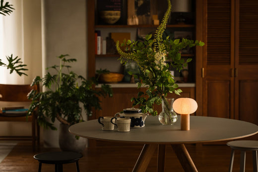 A gray table with a wooden edge. On the table is a table lamp "TORCHIN" made from Japanese Yame-chochin lanterns, a tea set, and a glass vase with a houseplant. The portable lamp is lit, emitting a warm lamp.